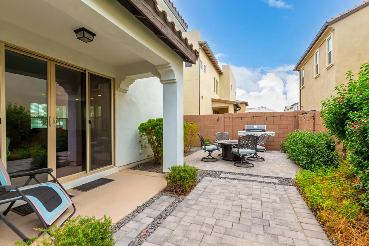 A cozy backyard patio featuring a comfortable seating area, a built-in grill, lush green shrubs, and sliding glass doors leading to the home. The setup is ideal for barbecues, outdoor dining, or simply relaxing with friends and family.