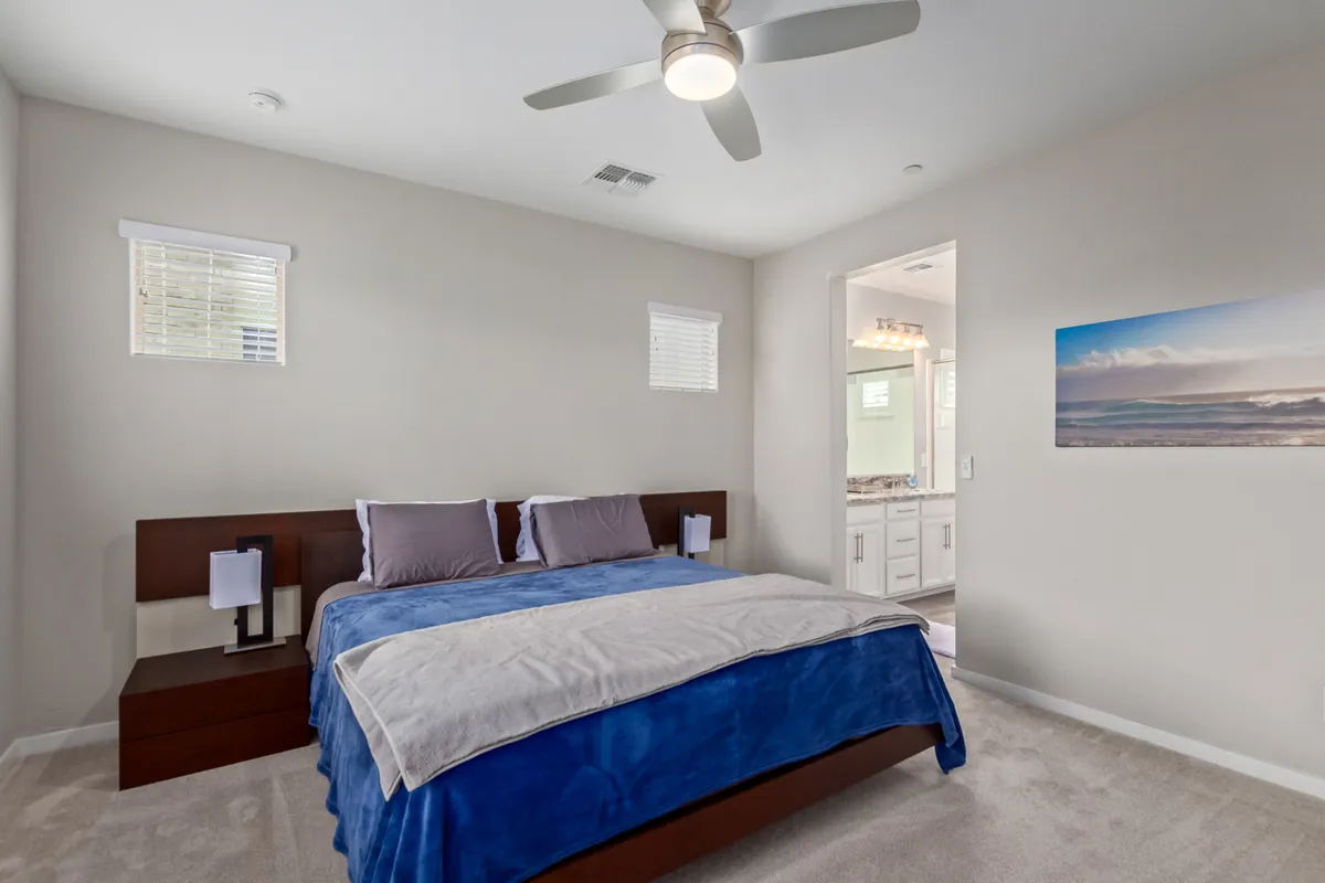 A tranquil bedroom featuring a bed with blue bedding, complemented by a pair of nightstands topped with lamps. Above the bed hangs wall art depicting a serene beach scene. Soft natural light fills the space, accentuating its airy and restful atmosphere.