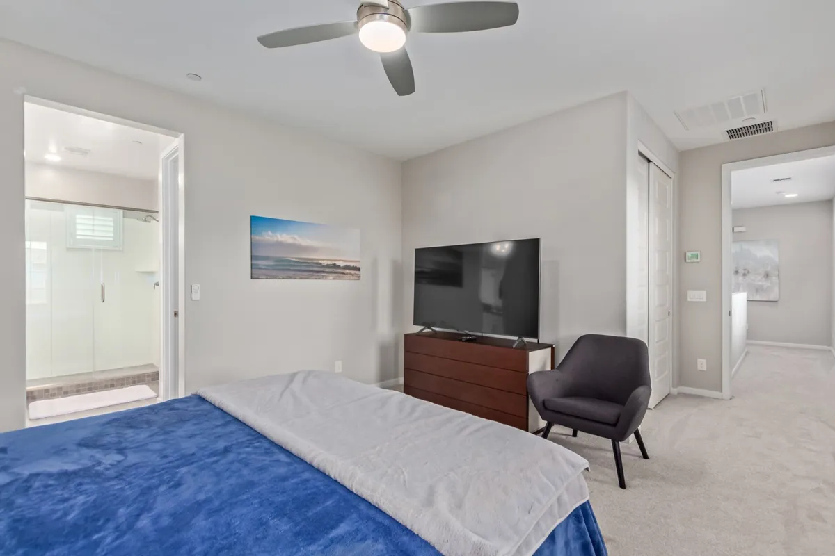 A spacious bedroom setup with a bed dressed in blue bedding, an armchair beside a large window, and a wall-mounted television. Artistic touches and abundant natural light give the space a welcoming and serene vibe, suitable for reading or relaxing.