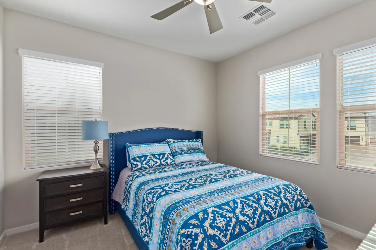 A cozy bedroom adorned with blue patterned bedding, a nightstand with a lamp, and a window with blinds. Sunlight streams through, creating a peaceful and inviting environment ideal for relaxing or reading.