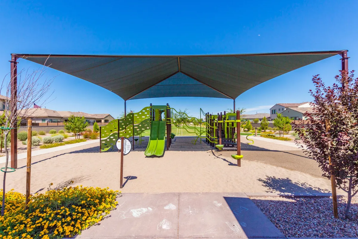 This image captures a vibrant playground scene featuring colorful playground equipment under a shade structure, surrounded by blooming flowers. Children are seen laughing and playing, while families gather around and enjoy the communal atmosphere.