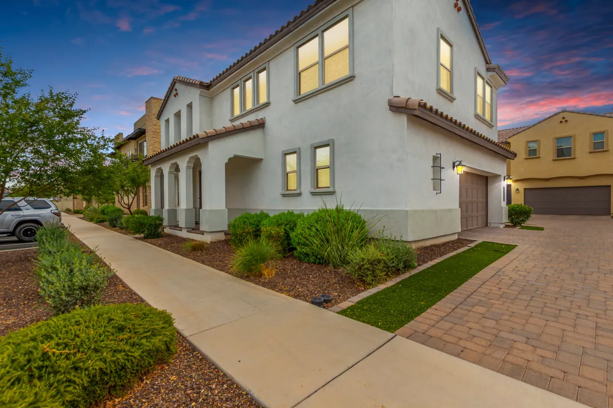 The image showcases the charming exterior of a spacious, two-story single-family house in Gilbert, Arizona. The inviting front porch is surrounded by a beautifully landscaped garden, with vibrant greenery and flowers, while a well-maintained driveway leads up to the home. The outdoor sitting area and the welcoming porch suggest a perfect spot for enjoying sunsets, quiet mornings, or hosting family gatherings.