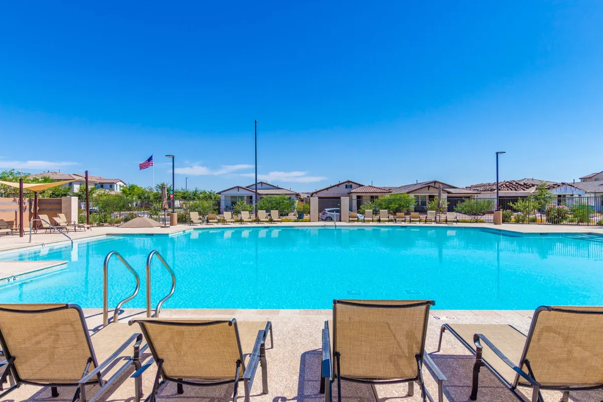 A sparkling community swimming pool bordered by lounge chairs and landscaped greenery. The pool sits within a tranquil neighborhood area, with residential buildings visible in the background. The setting suggests both relaxation and opportunities for casual socializing.