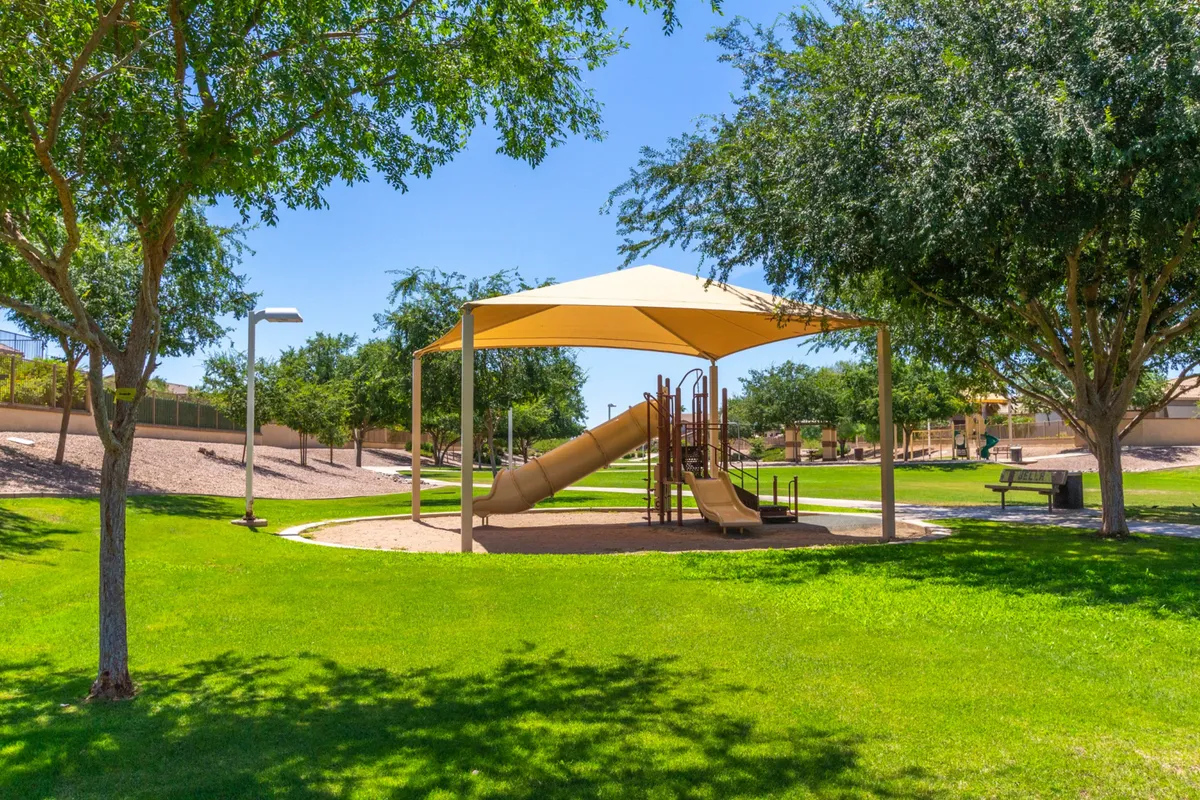 A lively playground set within a green park, complete with a shade structure and surrounded by mature trees. Children are seen playing, while families picnic and socialize, capturing a joyful, welcoming community vibe.